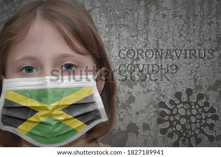Little girl in medical mask with flag of jamaica stands near the old vintage wall with text coronavirus, covid, and virus picture. Stop virus