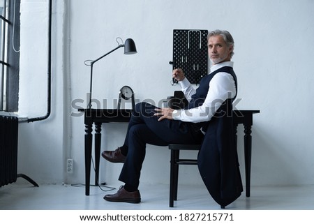 Pensive handsome matured male owner of corporation in elegant suit sitting at table while working in modern workplace.