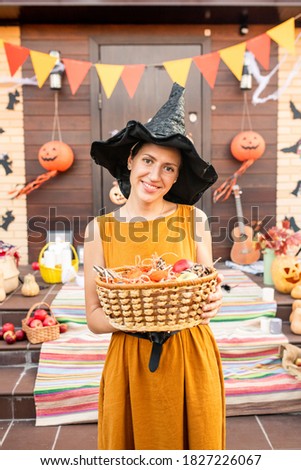 Young cheerful pretty female in black hat of halloween witch holding basket with sweets while waiting for halloween kids by decorated door