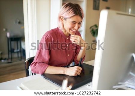 Indoor shot of cheerful happy young female with stylish pinkish hair laughing while working from home, sitting at desk with computer and graphic tablet, retouching images or drawing animation