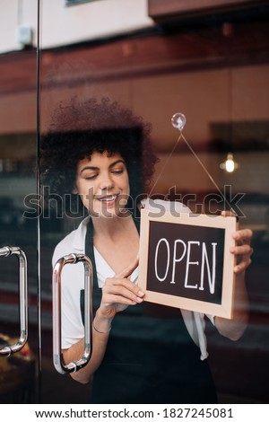 Portrait of beautiful business woman, with afro hairstyle, putting an open sign on the glass door. Happy female owner attending new customers. Small business concept, restaurants, birth of new store.