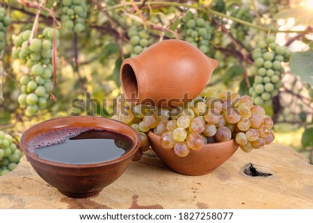Georgian traditional decorative jug for wine kvevri and clay cups with wine and grape on the wooden log slice, blurred vineyard background. Royalty-Free Stock Photo #1827258077