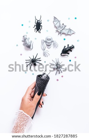Halloween card on white with black and silver holiday decor. Woman with black manicure holding black cone with bats and ghosts flying around, copy space, gothic holiday flat lay, vertical image