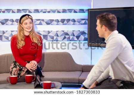 Two TV hosts talking to each other while sitting on a sofa in a film studio, they are in the process of filming a TV show.