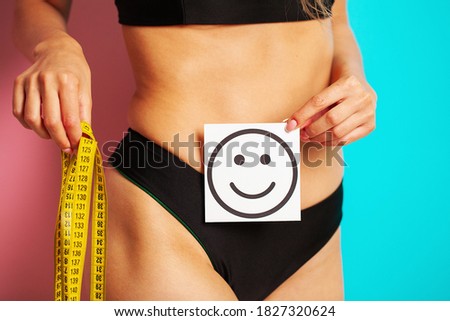 Close up of a woman with a slender figure shows the result holding a card near her belly with a smile and a yellow tape measure.
