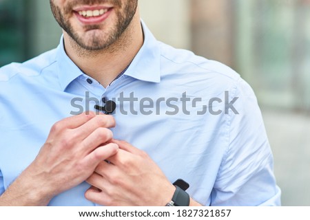 Young handsome man, male blogger adjusting small lavalier microphone on his shirt and smiling while standing outdoors