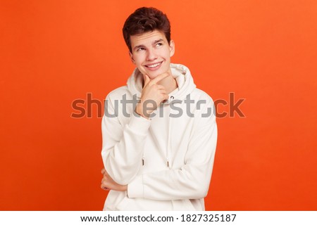Smiling young man with peaceful face thinking over plans for future holding his chin, thoughtful teenager dreaming about good job. Indoor studio shot isolated on orange background Royalty-Free Stock Photo #1827325187
