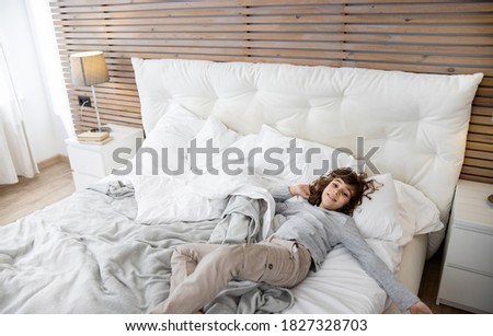 Handsome curly-haired boy lying and resting on the white bed at the modern bedroom. Stock photo