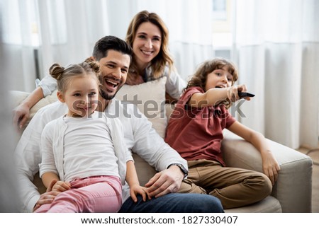 Leisure together. Happy family of four is enjoying at home. Small cute kids are rejoicing, parents are on the sofa, hugging, watching cartoons