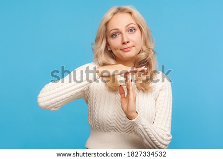 Serious blond woman in knitted sweater showing time out gesture, need more time for work , deadline, asking for pause. Indoor studio shot isolated on blue background Royalty-Free Stock Photo #1827334532