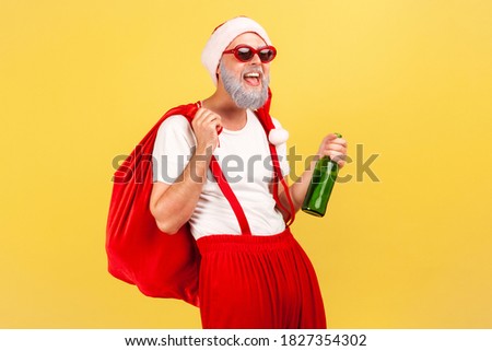 Happy elderly man in santa claus hat, trendy sunglasses and pants with suspenders holding bottle with alcohol and big red bag, celebration, bad habit. Indoor studio shot isolated on yellow background