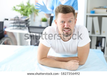 Young male patient lies on couch on stomach with doctor wearing rubber glove in background. Finger examination of rectum concept. Royalty-Free Stock Photo #1827356492