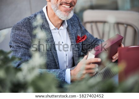 Cropped photo of an aged man smiling and holding a dark-red box while sitting in an outdoor cafe