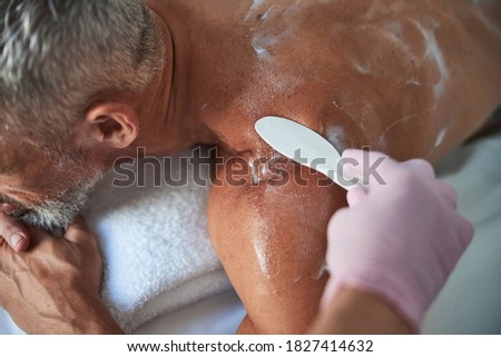 Cropped photo of spa worker holding a spatel while spreading lotion on aging man back