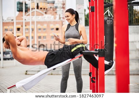 Jolly young female is taking photo on smartphone of strong guy doing abdominal crunches on bench in city