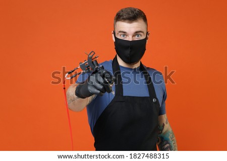 Young tattooer master artist man in blue t-shirt apron face mask hold in hand machine black ink jar equipment for making art body isolated on brown background studio. Tattoo translation life is fight.
