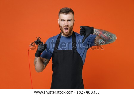 Displeased tattooer master artist tattooed man in apron hold machine black ink in jar equipment for making art on body showing thumb down isolated on brown background. Tattoo translation life is fight