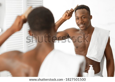 Shirtless Black Guy Brushing And Combing Hair Doing Haistyling Using Hairbrush Standing Near Mirror In Modern Bathroom At Home. Male Haircare Routine. Selective Focus