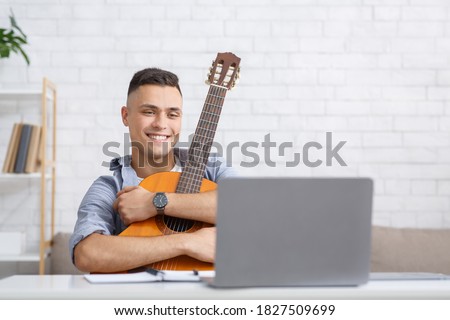 New hobby during self-isolation, app for music education and guitar playing. Happy man hugs guitar and looks at laptop, watch online lesson at home interior during coronavirus epidemic, free space