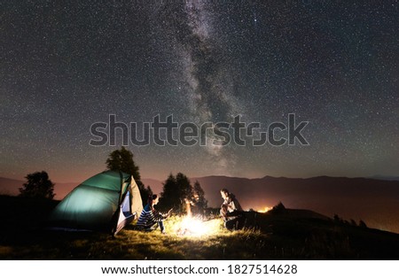 Young romantic couple hikers having a rest at campfire beside camp and tourist tent under night sky full of stars and Milky way. On the background beautiful starry sky, mountains and luminous town