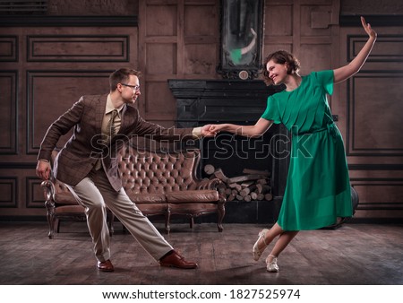 a young couple dancing swing in a retro hall in front of a fireplace and a leathern sofa; the woman wears a green dress Royalty-Free Stock Photo #1827525974