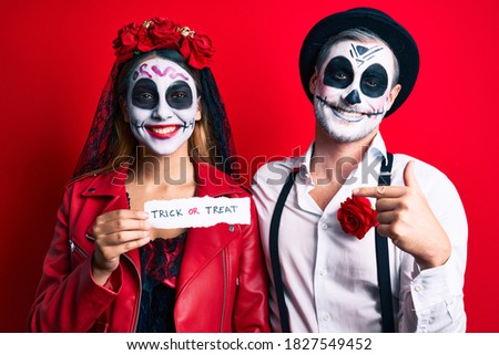 Couple wearing day of the dead costume holding trick or treat paper pointing finger to one self smiling happy and proud 