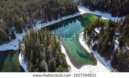 A top down drone shot of Green Lake in Austrian Alps. The lake shimmers with many shades of green and turquoise. Thick forest around it. Winter in the mountains. There is snow on the ground. Wandering