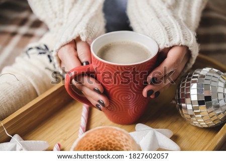 Young woman sits on plaid in cozy knitted woolen white sweater and holds cup of cocoa in her hands. Hygge wooden tray with mug of chocolate, toy tree, candle, stars.