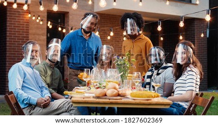 Portrait of mixed-races people in face shields at dinner table in back yard and smiling to camera. Multi ethnic family at picnic in court outdoors. Pandemic communication of generations. Royalty-Free Stock Photo #1827602369