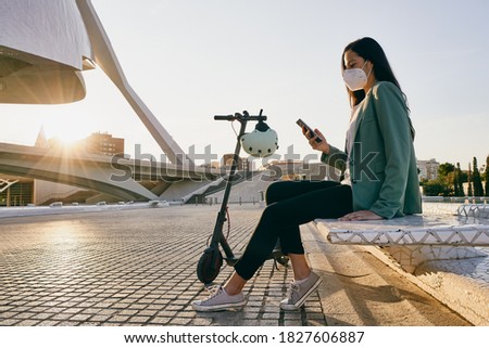 Young woman sitting on a bench using a mobile phone. Woman wearing face mask while using an electric scooter                