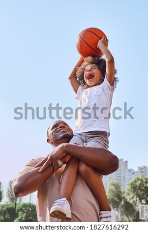Happy African American father lifting his kid up and helping him score a basketball into a hoop