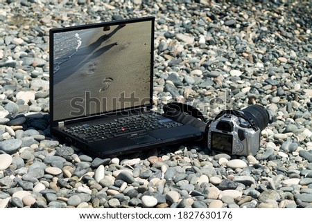Laptop with picture of footprints on sand on its monitor and camera laying among gray pebbles on sea beach