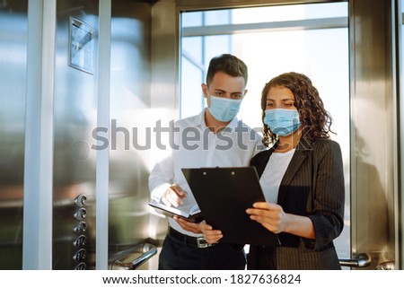 Business partners in protective sterile mask in the elevator. Colleagues talking while standing in elevator at  modern office. Teamwork during pandemic in quarantine city. Covid-19. Royalty-Free Stock Photo #1827636824