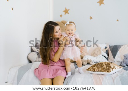 A young mother plays with her one-year-old daughter, sitting on the bed in the children's room. They are happy and smiling