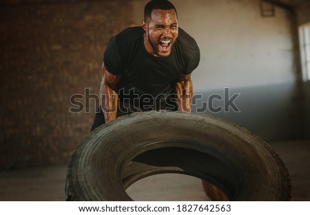 Male athlete flipping heavy tire inside an abandoned warehouse. Strong man flipping a tyre during an intense training session in a cross workout space. Royalty-Free Stock Photo #1827642563