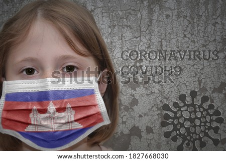 Little girl in medical mask with flag of cambodia stands near the old vintage wall with text coronavirus, covid, and virus picture. Stop virus