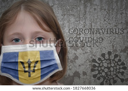 Little girl in medical mask with flag of barbados stands near the old vintage wall with text coronavirus, covid, and virus picture. Stop virus