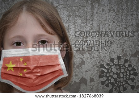 Little girl in medical mask with flag of china stands near the old vintage wall with text coronavirus, covid, and virus picture. Stop virus