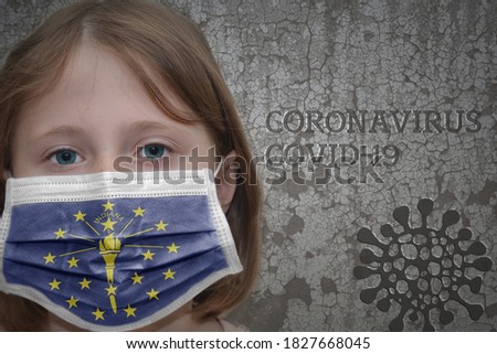 Little girl in medical mask with indiana state flag stands near the old vintage wall with text coronavirus, covid, and virus picture. Stop virus