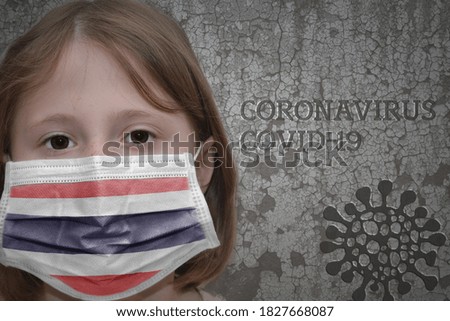 Little girl in medical mask with flag of thailand stands near the old vintage wall with text coronavirus, covid, and virus picture. Stop virus
