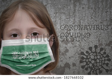 Little girl in medical mask with flag of saudi arabia stands near the old vintage wall with text coronavirus, covid, and virus picture. Stop virus