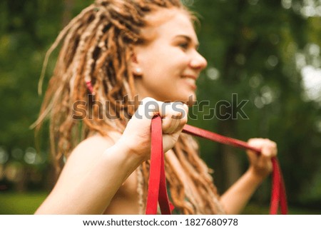 Sporty smiling young woman with dreadlocks training in the park with red elastic band in sportswear. blured background