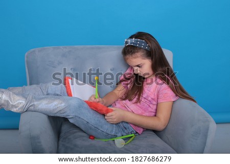 An eight-year-old girl with dark brown hair sits on a blue armchair and draws in an orange diary