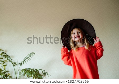 little girl in a witch hat with an orange ball in her hand on a yellow background. Halloween concept.Active games at home. Laughs emotionally and jumps on the bed.