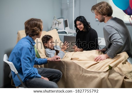 A little cheerful girl showing something on her tablet to her elder brother and her mom, the girl is staying in a hospital bed, the family are visiting.
