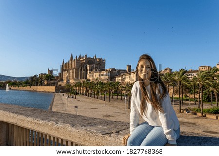 Young woman with light skin and brown hair sitting in front of the cathedral of Palma in autumn smiling at the camera, wears a white sweater and striped pants