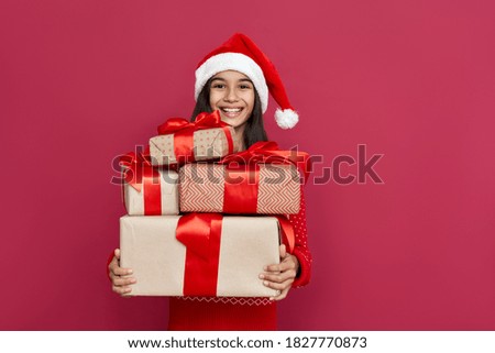 Funny indian latin preteen girl wears santa hat looking at camera holding many gifts boxes standing isolated on red background. Merry Christmas presents shopping sale, Happy 2022 New Year celebration. Royalty-Free Stock Photo #1827770873