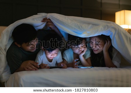 Asian happy family mother, father and little daughter and son watching movie or cartoon in smartphone together and blanket cover their head in bed at night at home
