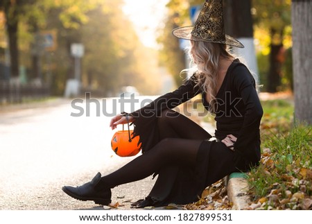 Halloween Witch with pumpkin. Beautiful young woman in a witch costume on the street. Halloween decor. Copy space.