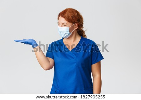 Medical workers, covid-19 pandemic, coronavirus concept. Smiling pleasant female doctor, nurse looking happy as holding something on hand, wearing face mask and rubber gloves